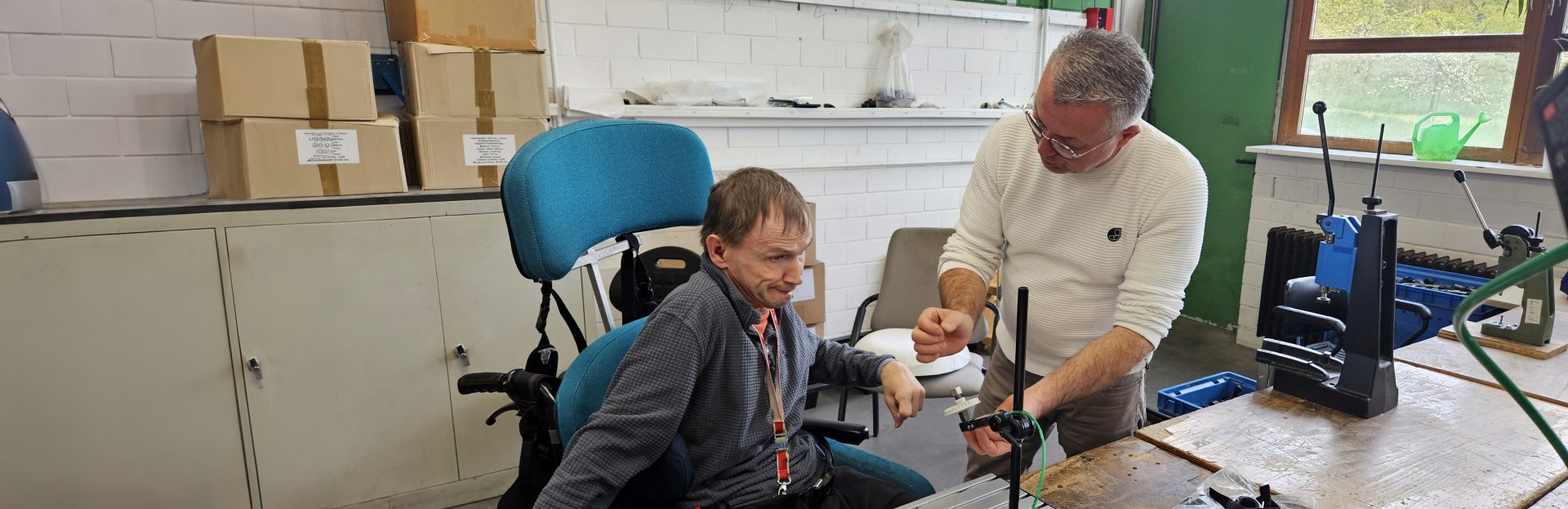 Zwei Personen in einer Werkstatt. Ein Mann sitzt im Rollstuhl. Der andere Mann steht rechts daneben und erklärt eine Maschine.