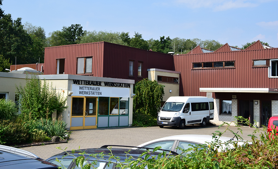 Die Wetterauer Werkstätten sind ein zweistöckiges braunes Gebäude. Links ist der Eingang zu sehen. Vor dem Haus steht ein weißer Transporter.