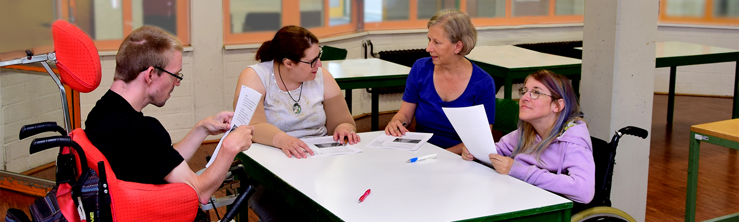 Ein Mann und drei Frauen sitzen im Büro für Leichte Sprache an einem Tisch und unterhalten sich über einen Text.