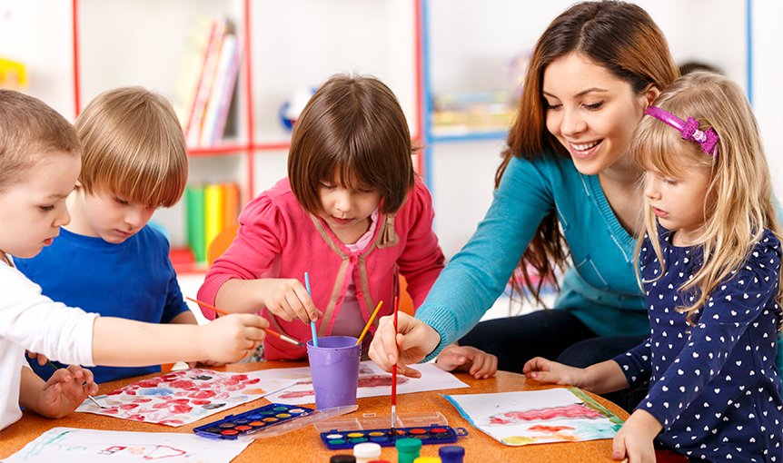 In der Kita sitzen drei Kinder und eine Erzieherin am Tisch und malen mit Wasserfarben.