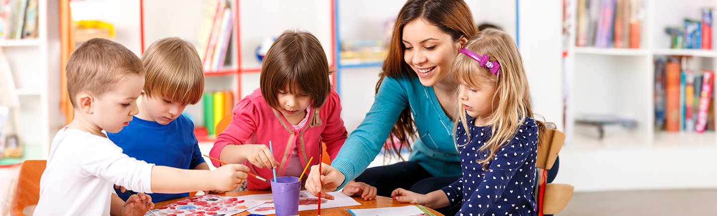 In der Kita sitzen drei Kinder und eine Erzieherin am Tisch und malen mit Wasserfarben.