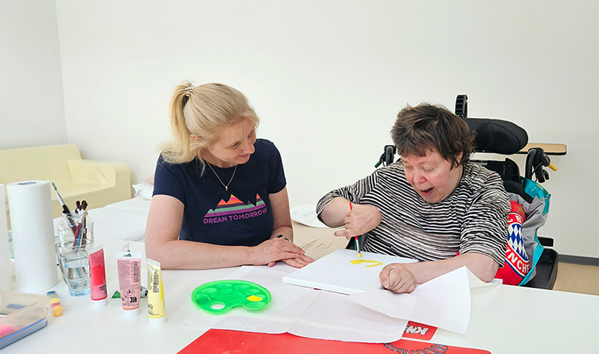 In der Tagesförderstätte sitzen zwei Frauen sitzen an einem Tisch. Eine Frau sitzt im Rollstuhl und malt mit einem Pinsel ein Bild. Die andere Frau leitet sie an.