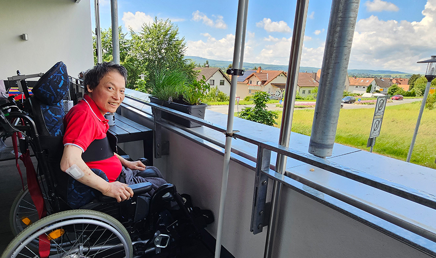 Verhinderungspflege. Ein Mann im Rollstuhl sitzt auf einem Balkon mit Aussicht ins Grüne.