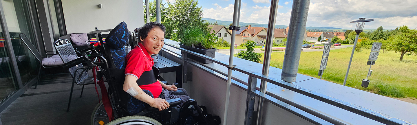 Verhinderungspflege. Ein Mann im Rollstuhl sitzt auf einem Balkon mit Aussicht ins Grüne.