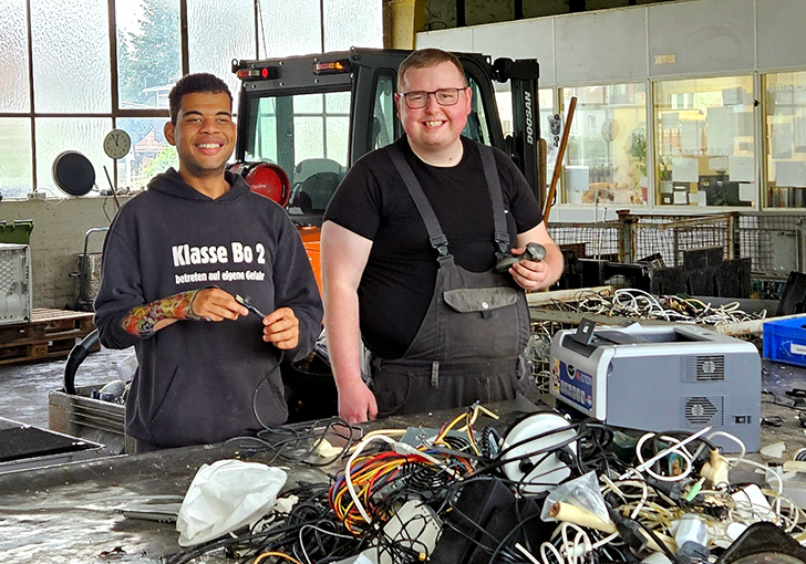 Zwei junge Männer in Arbeitskleidung stehen nebeneinander an einer Werkbank. Vor ihnen liegen viele Kabel.