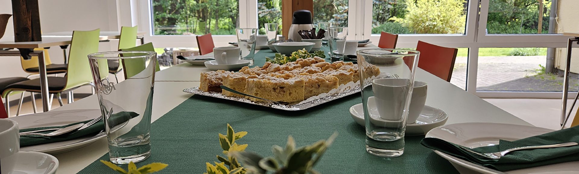 Ein für das Trösterkaffee gedeckter Tisch mit eine dunkelgrünen Tischdecke, weißem Geschirr und Gläsern. In der Mitte vom Tisch steht eine Platte mit Streuselkuchen.