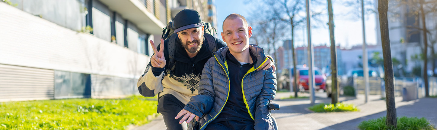 Zwei Männer sind auf einem Gehweg, einer sitzt im Rollstuhl, der andere beugt sich herunter und zeigt das Victory-Zeichen. Er macht ein Duales Studium.