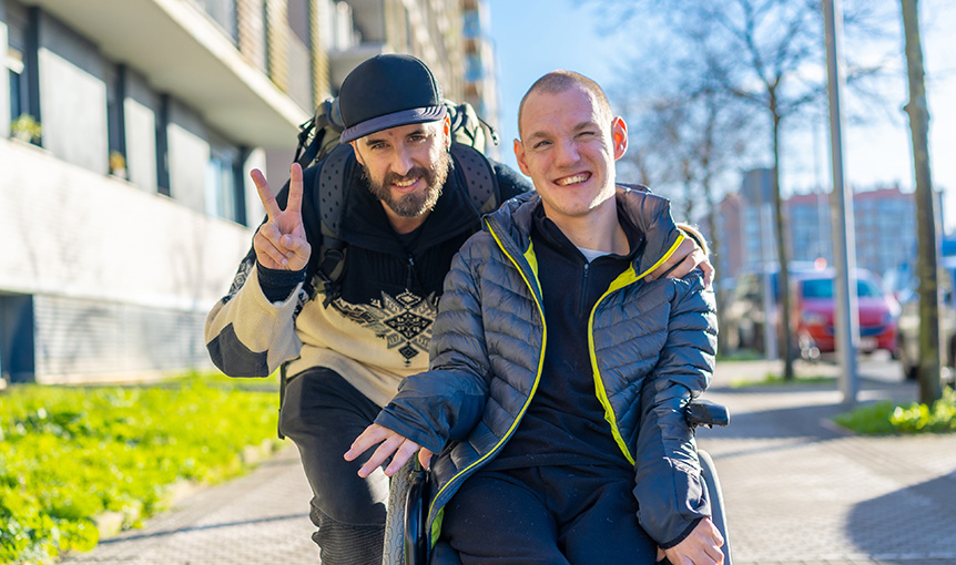 Zwei Männer sind auf einem Gehweg, einer sitzt im Rollstuhl, der andere beugt sich herunter und zeigt das Victory-Zeichen. Er macht ein Duales Studium.