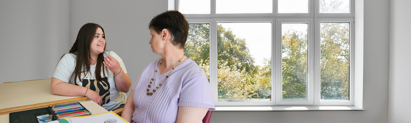 Zwei erwachsene Frauen sitzen in einem Raum am Tisch. Sie schauen sich an und sprechen miteinander. Die Frau auf der linken Seite hat eine Ausbildung zur Heilerziehungspflegerin gemacht.