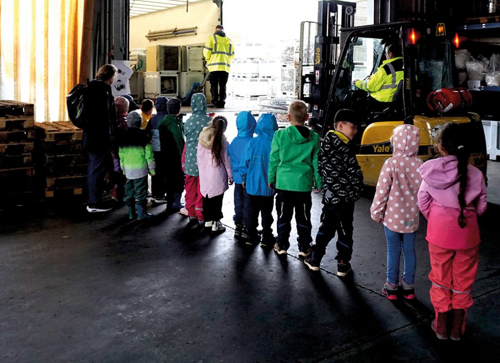 Eine Reihe von Kindern in bunter Regenkleidung steht mit dem Rücken zur Kamera. Die Kinder schauen in einen LKW, in dem alte Elektrogeräte gestapelt sind. Im Hintergrund ist ein Gabelstapler.