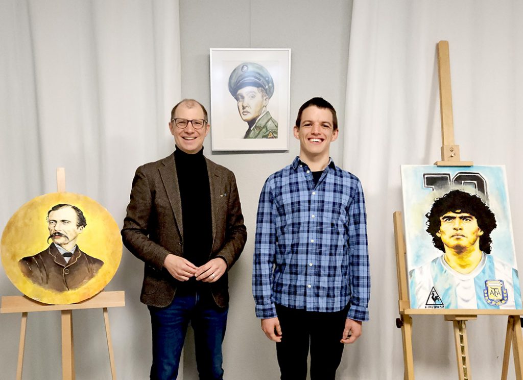 In einem Raum mit weißen Wänden stehen zwei Männer: links Jan Weckler, Landrat vom Wetteraukreis, rechts Viktor Lekov. Neben ihnen sind drei Porträts zu sehen. Die Kunstwerke gehören zur Ausstellung Viktor Art.