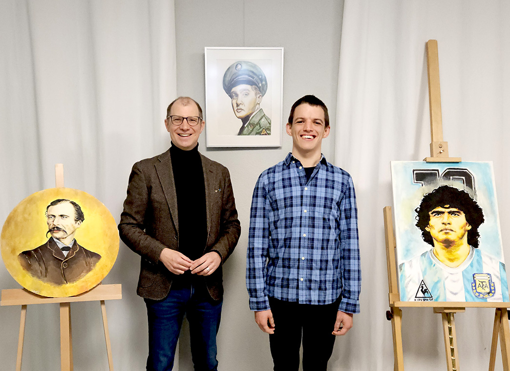In einem Raum mit weißen Wänden stehen zwei Männer: links Jan Weckler, Landrat vom Wetteraukreis, rechts Viktor Lekov. Neben ihnen sind drei Porträts zu sehen. Die Kunstwerke gehören zur Ausstellung Viktor Art.