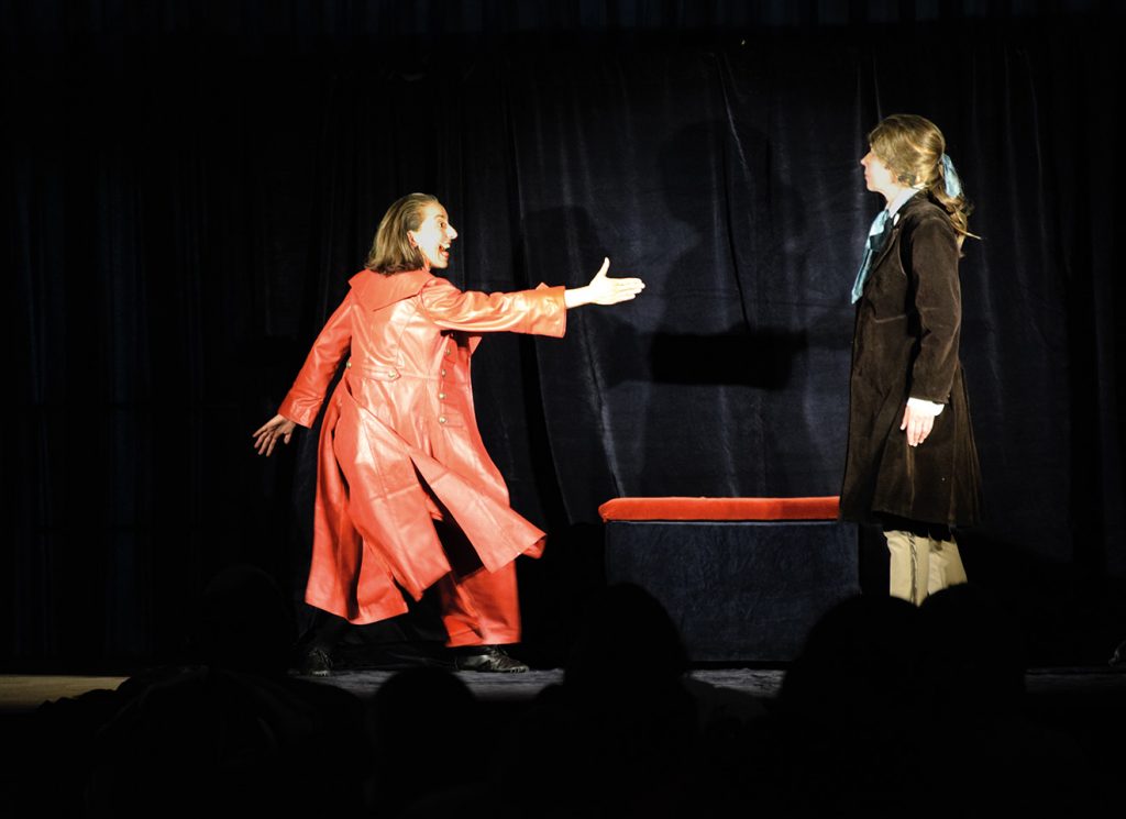 im Theater: Eine Frau in einem roten Mantel spielt den Teufel. Sie streckt den Arm aus und hält die Hand zum Einschlagen hin. Rechts steht Faust mit einem braunen Gehrock. Er schaut zum Teufel.