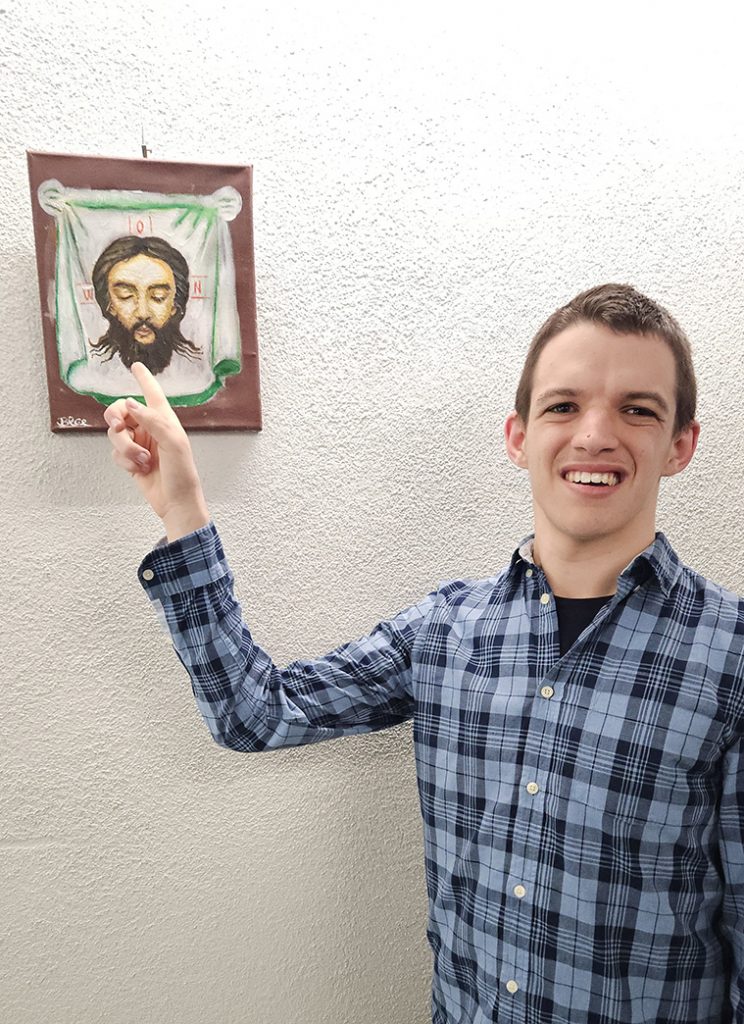 Viktor Lekov, schaut in die Kamera und lacht. Er zeigt auf ein Kunstwerk, das er in der Ausstellung Viktor Art präsentiert.