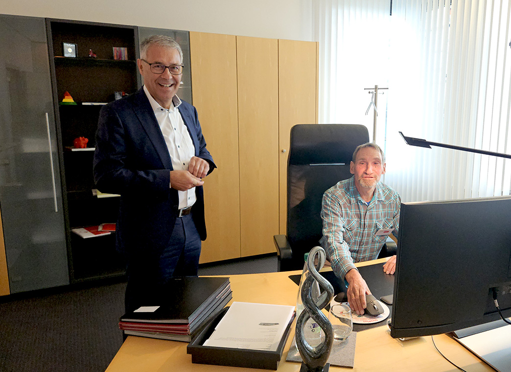 Ein Mann steht neben einem Schreibtisch, an dem ein zweiter Mann vor einem Computer sitzt.