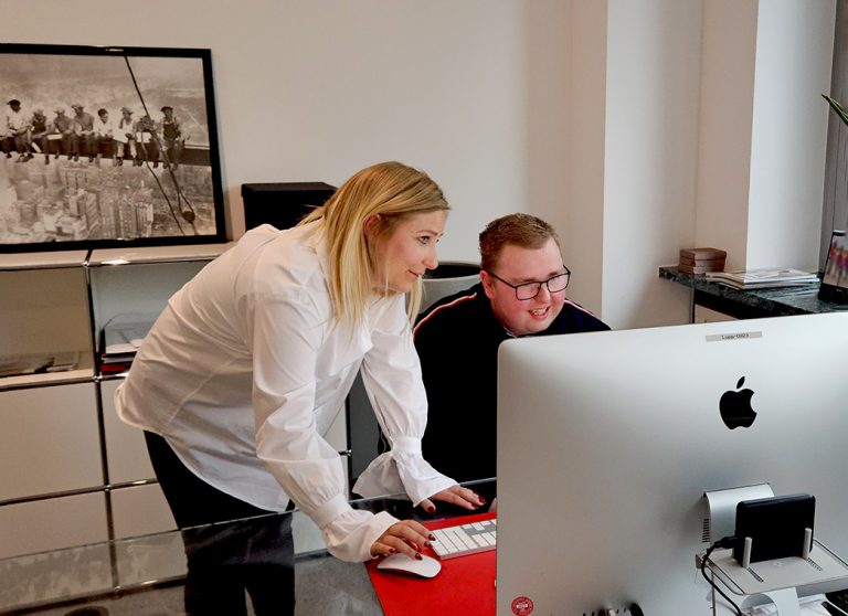 Eine Frau steht neben einem Mann, der am Schreibtisch sitzt, und zeigt ihm etwas am Computer.