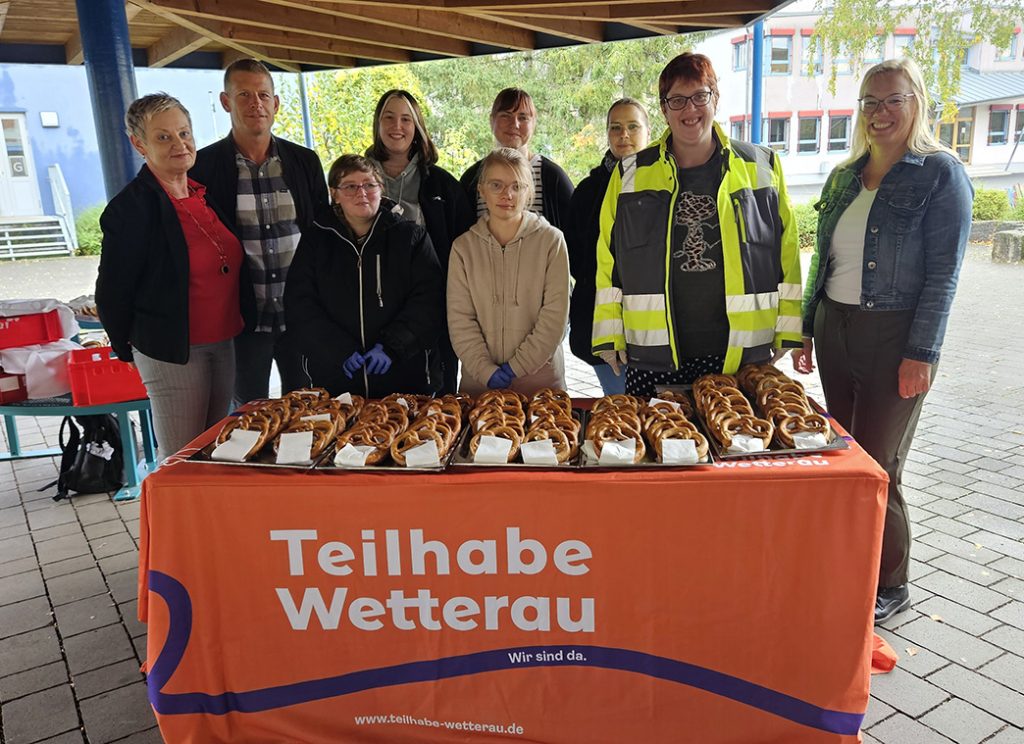 Brezelaktion im Gymnasium Nidda: Eine Gruppe von Menschen steht hinter einem Tisch mit einer orangenen Tischdecke, auf der Teilhabe Wetterau steht. Auf dem Tisch liegen frisch gebackene Laugenbrezeln.