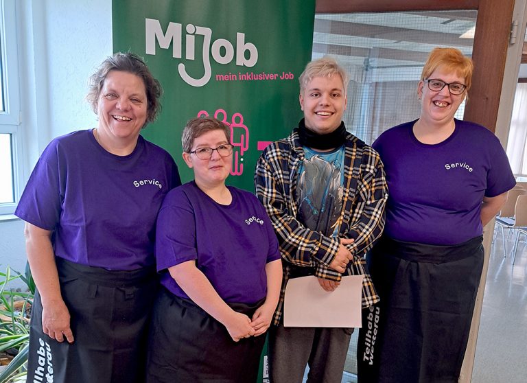 Drei Frauen mit lila T-Shirts und schwarzen Schürzen und ein junger Mann stehen nebeneinander und lachen in die Kamera.