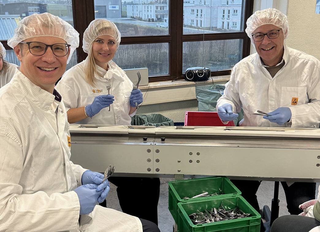 Drei Personen mit Kittel, Haarnetz und Handschuhen sitzen an einer Maschine. Sie halten Besteck in den Händen.