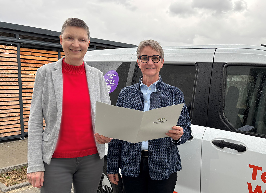 Zwei Frauen stehen nebeneinander vor einem weißen Auto. Sie halten eine weiße Mappe. Beide lächeln in die Kamera.