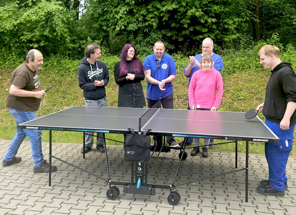 Zwei Männer spielen im Freien Tischtennis. Drei Männer und zwei Frauen stehen hinter der Tischtennisplatte und klatschen.