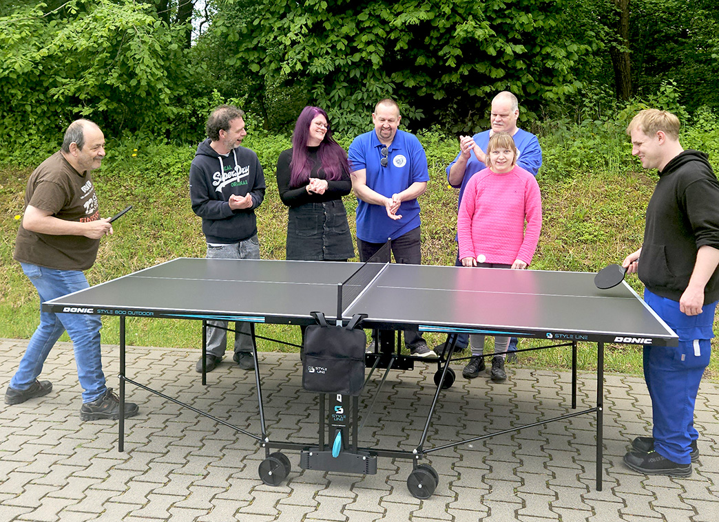 Zwei Männer spielen im Freien Tischtennis. Drei Männer und zwei Frauen stehen hinter der Tischtennisplatte und klatschen.